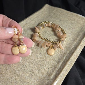 Puede incluir: Un conjunto de joyas de color melocotón, que incluye pendientes y una pulsera. Los pendientes presentan pequeñas piedras y colgantes de conchas marinas. La pulsera tiene una banda dorada con cuentas con virutas de piedra de color melocotón y un dije de concha.