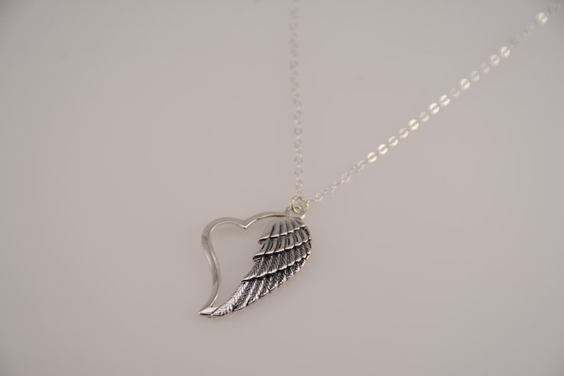 May include: A silver necklace with a heart-shaped pendant featuring a detailed angel wing design.