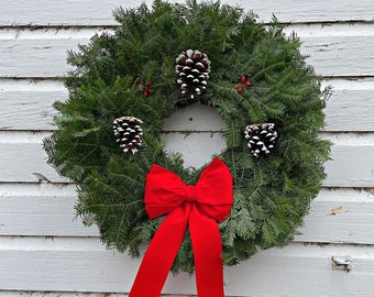 May include: A fresh evergreen wreath with three pinecones and a bright red bow. The wreath is decorated with small red berries and is displayed against a white, weathered wooden background.