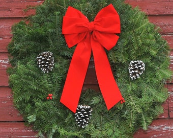 May include: A circular evergreen wreath made of dense green pine needles, featuring a large, bright red fabric bow at the top center. Three pine cones with white-tipped scales are arranged around the wreath, accented by small red berries.