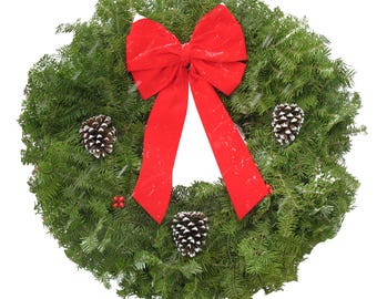 May include: A circular evergreen wreath featuring dense green pine needles, a large bright red fabric bow at the top, three brown pinecones with white-tipped scales, and small clusters of red berries nestled within the foliage.