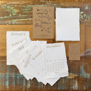 May include: A collection of handmade paper calendar pages and informational cards. The calendar pages are white with brown text, displaying months and dates for 2026. A brown card features text: "A tapestry of repurposed materials blended together to create..."