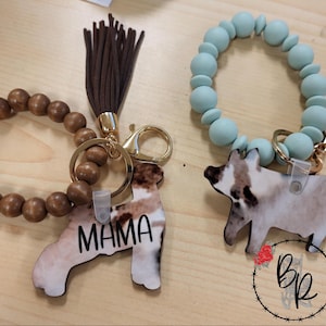 May include: Two wristlet keychains. One features a brown wooden bead bracelet with a brown tassel and a gold-tone clasp. The other is a light blue bead bracelet. Both have a pig-shaped charm. One charm says "MAMA".
