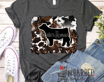 May include: A dark gray t-shirt with a black silhouette of a cow on a brown, white, and black animal print background. The text "The Barbed Rocker" is visible in the bottom right corner of the image.