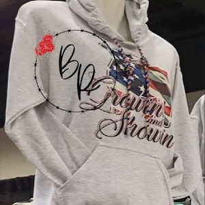 May include: A light gray hooded sweatshirt with a front pocket and drawstring. The sweatshirt features a graphic of the American flag and the words "Grown and Shown" in a script font. A circular design with the letters "BP" and red roses is on the left side.