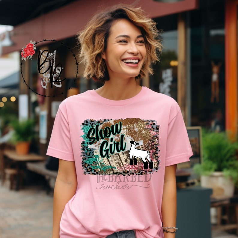 May include: Pink t-shirt with a graphic design featuring a white lamb, a wooden plank, and the text "Show Girl" and "The Barbed Rocker".
