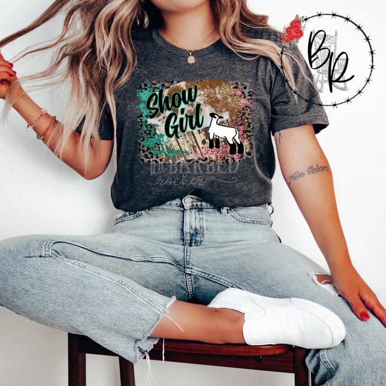 May include: A gray t-shirt with a graphic design featuring a white lamb, a wooden fence, and the text "Show Girl". The graphic is surrounded by a colorful, glittery background.