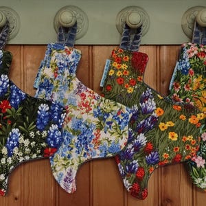 May include: Four Texas-shaped pot holders with different floral patterns. The pot holders are made of fabric and have a loop for hanging. The patterns include bluebonnets, wildflowers, and other floral designs.