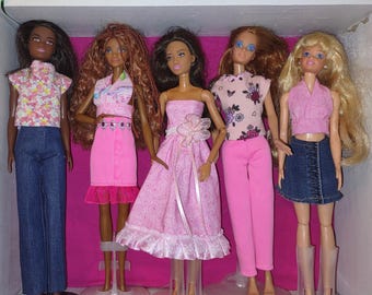 May include: Five fashion dolls standing in a row. The first wears a floral top and blue jeans. The second has long curly hair, a pink top, and a pink skirt. The third wears a pink dress with a floral accent. The fourth has a floral top and pink pants. The fifth has blonde hair, a pink top, and a blue denim skirt.