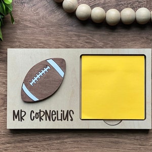May include: A wooden desk organizer with a cutout for a sticky note pad. The organizer has a brown football design and the text "MR CORNELIUS" on the front.