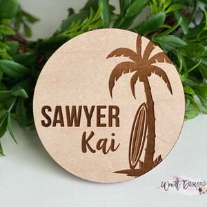 May include: A round wooden sign with a palm tree and surfboard design. The sign says "Sawyer Kai" in brown lettering.