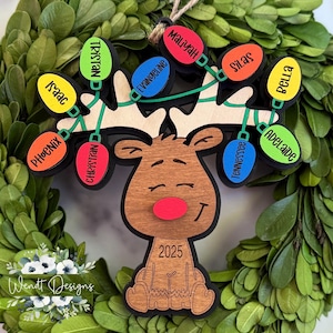 May include: A wooden reindeer Christmas ornament with a red nose and a string of colorful lights, each bearing a name. The ornament is set against a green wreath and includes the year 2025.