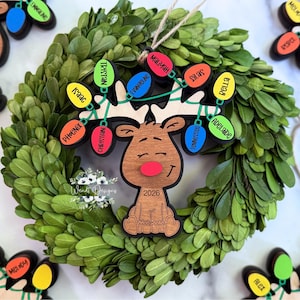 May include: A wooden reindeer Christmas ornament featuring a red nose and the year 2026. The antlers are decorated with colourful light bulb shapes, each engraved with names including Tristan, Isaac, Phoenix, Christian, Evangeline, Maliyah, Silas, Bella, and Adelaide.