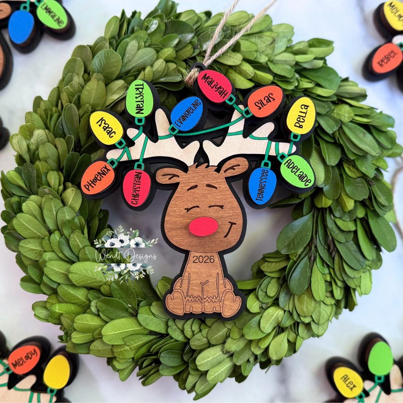 May include: A wooden reindeer Christmas ornament featuring a red nose and the year 2026. The antlers are decorated with colourful light bulb shapes, each engraved with names including Tristan, Isaac, Phoenix, Christian, Evangeline, Maliyah, Silas, Bella, and Adelaide.