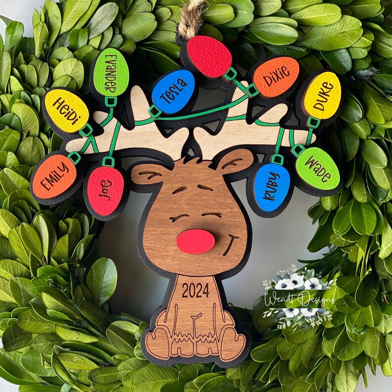 May include: A wooden reindeer ornament featuring a red nose and the year 2024 engraved on its chest. The reindeer has antlers decorated with a string of colourful light bulb-shaped wooden tags, each personalised with names including Evander, Tesla, Dixie, Duke, Wade, Ruby, Joe, Heidi, and Emily.