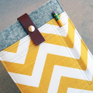 Puede incluir: Una bolsa de cremallera de fieltro gris con un patrón de chevron amarillo y blanco. La bolsa tiene una correa de cuero marrón con una hebilla de metal y un bolsillo para un lápiz.