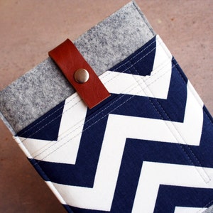 Puede incluir: Una funda de fieltro gris con un patrón de chevrón azul marino y blanco. Cuenta con una correa de cuero marrón y un cierre de botón a presión plateado. Diseñada para guardar una tableta o un lector de libros electrónicos.