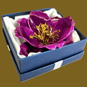 May include: A purple flower brooch with gold accents, displayed in a blue velvet box with white satin lining.