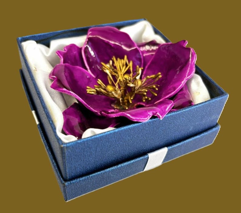 May include: A purple flower brooch with gold accents, displayed in a blue velvet box with white satin lining.