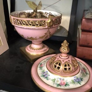 Könnte beinhalten: Eine rosafarbene Porzellanurne mit goldener Verzierung und floralem Design. Die Urne hat einen Deckel mit einem dekorativen Knauf und einem durchbrochenen Muster. Die Urne ist mit getrocknetem Pflanzenmaterial gefüllt.