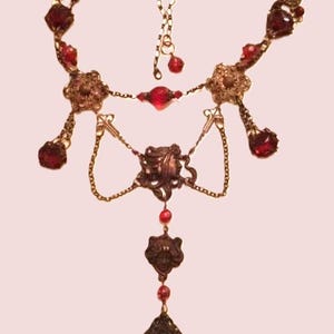 Könnte beinhalten: Eine lange, mehrreihige Halskette mit einem dunkelroten und goldenen Design. Die Halskette verfügt über mehrere Ketten, verzierte Metallakzente und dunkelrote Glasperlen.