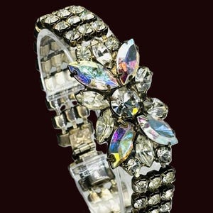 Op de afbeelding: Een vintage zilverkleurige armband met heldere, sprankelende strass-steentjes. De armband heeft een bloemmotief met iriserende, ovaalvormige stenen in het midden. De band bestaat uit meerdere rijen kleine, vierkante strass-steentjes.