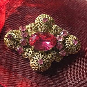 May include: A gold-toned brooch with a large pink oval rhinestone in the center. The brooch is decorated with smaller pink rhinestones and intricate filigree work.