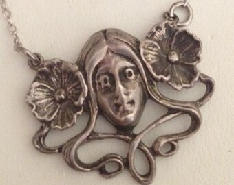 May include: Silver pendant necklace with a stylized face and floral details. The pendant hangs from a fine chain. The design has an Art Nouveau aesthetic. The necklace is made of silver.