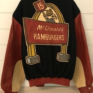 Puede incluir: Una chaqueta de béisbol vintage negra y roja con un gran parche bordado en la espalda. El parche presenta a un chef de dibujos animados sosteniendo un cartel que dice "15c" y un cartel que dice "McDonald's Hamburgers".