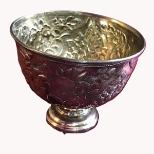 May include: A silver-coloured bowl with a decorative floral pattern. The bowl has a wide, angled rim and sits on a small, round base. The interior of the bowl has a gold hue.