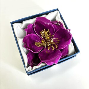 May include: A large, purple flower brooch with a gold centre and a detailed design. The brooch is in a blue box with white lining.