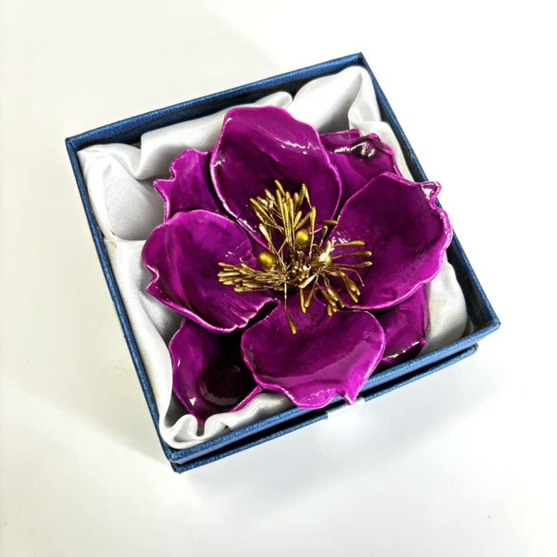 May include: A large, purple flower brooch with a gold centre and a detailed design. The brooch is in a blue box with white lining.