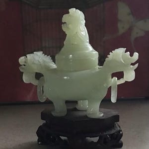 May include: A pale green jade incense burner with a detailed design. The burner features a mythical creature design with two dragon-like figures on either side. It sits on a dark brown wooden stand, showcasing intricate carvings. The background is a blurred red.