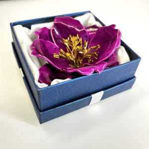 May include: A purple flower brooch with gold accents in a blue box with white lining.