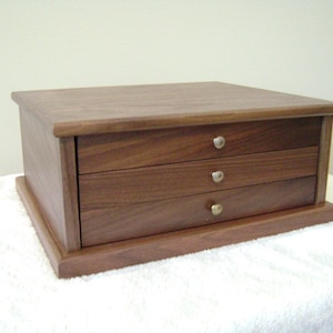 May include: A small, wooden jewelry box with three drawers. The box is made of dark brown wood and has a rectangular shape. Each drawer has a small, round, silver-colored knob. The box sits on a slightly raised base.