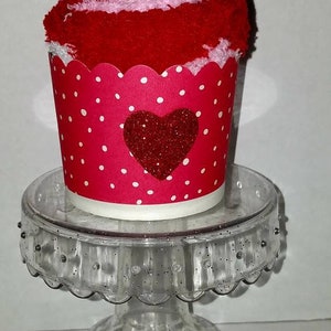 May include: A red and white sock cupcake with a red glitter heart on a clear cupcake stand.