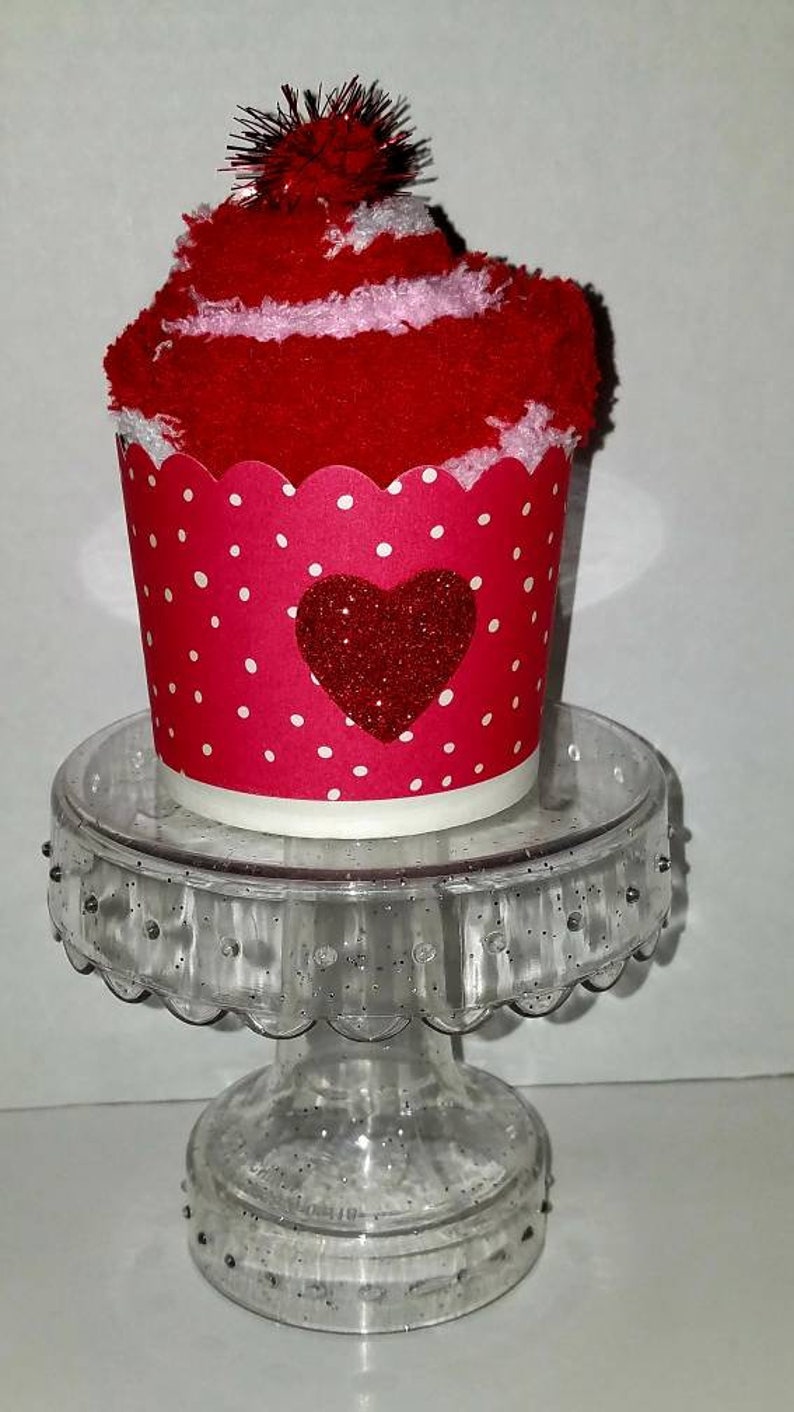 May include: A red and white sock cupcake with a red glitter heart on a clear cupcake stand.