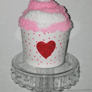 May include: A pink and white cupcake sock with a red glitter heart and a red pom-pom on top. The cupcake sock is sitting on a clear glass pedestal.
