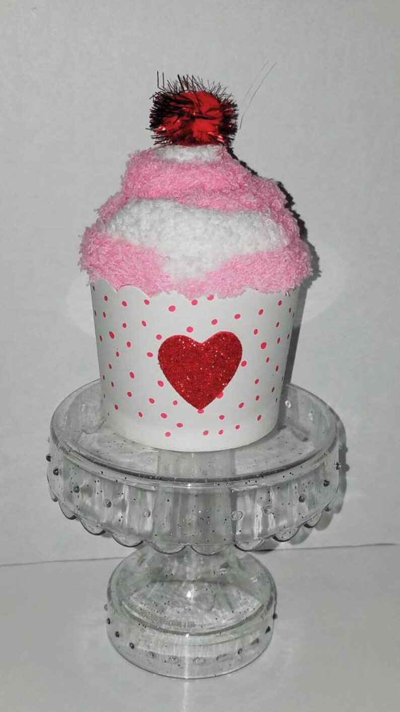 May include: A pink and white cupcake sock with a red glitter heart and a red pom-pom on top. The cupcake sock is sitting on a clear glass pedestal.