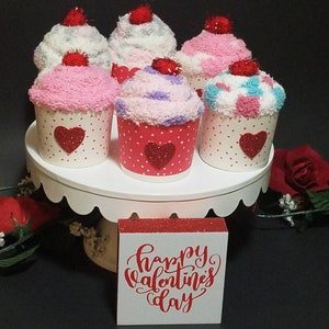 May include: Six sock cupcakes decorated with red glitter hearts and white polka dots. The cupcakes are arranged on a white cake stand. A small wooden sign with red glitter lettering reads "Happy Valentine's Day".