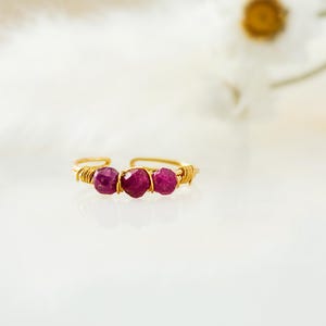 May include: A gold-toned wire ring featuring three faceted, deep red gemstones. The gemstones are centered on the ring, with the wire wrapped around them. The ring is set against a soft, white background, with a blurred flower in the background.