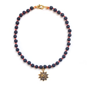 May include: A necklace featuring dark blue lava rock beads strung on red cord. A gold-colored sunburst pendant hangs at the center. The necklace is secured with a gold-colored clasp.
