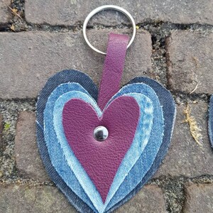 Op de afbeelding: Een hartvormige sleutelhanger gemaakt van denim en leer. Het hart is gelaagd met verschillende tinten blauw denim en heeft een bordeauxrood leren middenstuk met een zilveren knoop.