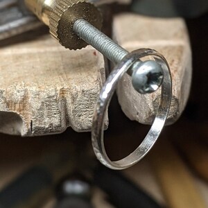 Op de afbeelding: Close-up van een zilveren ring die op zijn plaats wordt gehouden door een metalen klem op een houten oppervlak. De ring heeft een getextureerd oppervlak en is gebogen. De klem heeft een messing fitting en een schroef. De achtergrond is wazig.