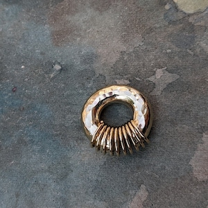 Puede incluir: Un colgante de anillo dorado con textura martillada y una serie de anillos m&aacute;s peque&ntilde;os apilados dentro del anillo m&aacute;s grande.