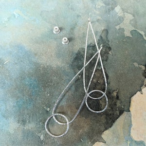 Op de afbeelding: Zilverkleurige oorhangers met een gehamerde textuur, met een druppelvorm en twee verbonden cirkels. Twee oorbelruggen zijn ook zichtbaar. De oorbellen worden gepresenteerd op een aquarelachtergrond met tinten blauw, groen en perzik.