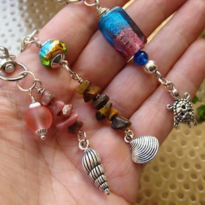 Op de afbeelding: Handgemaakte sleutelhangers met zeethema-bedels. Elke sleutelhanger heeft een unieke combinatie van kralen, waaronder gekleurd glas, tijgeroog en roze stenen. Bedels zijn onder meer een zeeschelp, een schildpad en een zeeslak.