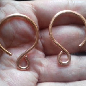 May include: Two copper-colored, handmade ear cuffs with a unique, open design. The ear cuffs have a curved top and a looped bottom. A dark ring is also visible.