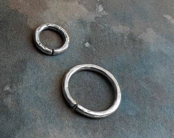 May include: Two silver-colored, hammered-texture rings. One ring is smaller, and the other is larger. Both rings have a small opening. The rings are likely jewelry, such as earrings or nose rings.