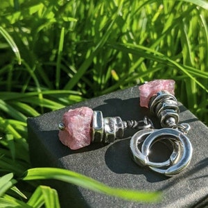 May include: A pair of silver earrings with pink gemstone accents. The earrings are set on a black box, surrounded by green grass. The earrings have a unique design, with a combination of metal and rough-cut stones.